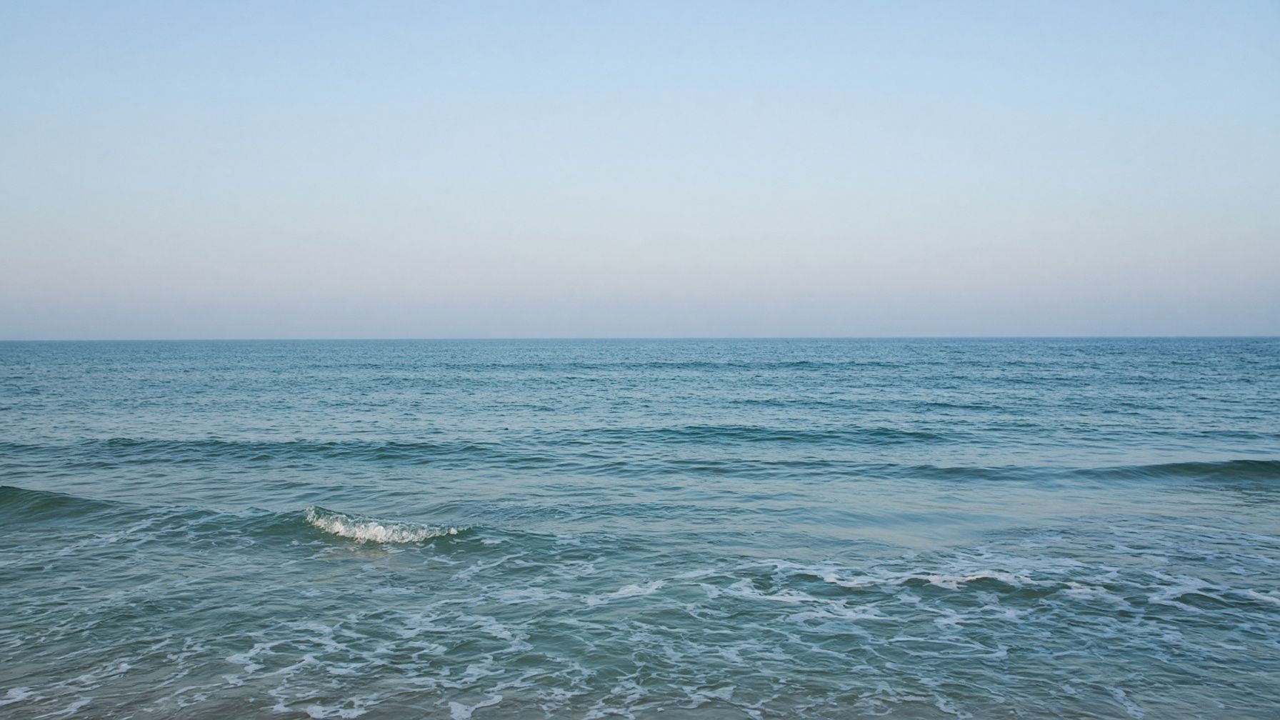 A quiet ocean shoreline at dusk. Wet sand in the foreground. Empty sky over the water.